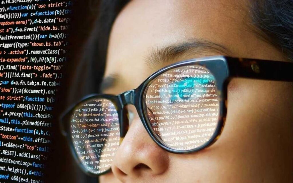 A screen with code is reflected in the glasses of a young person