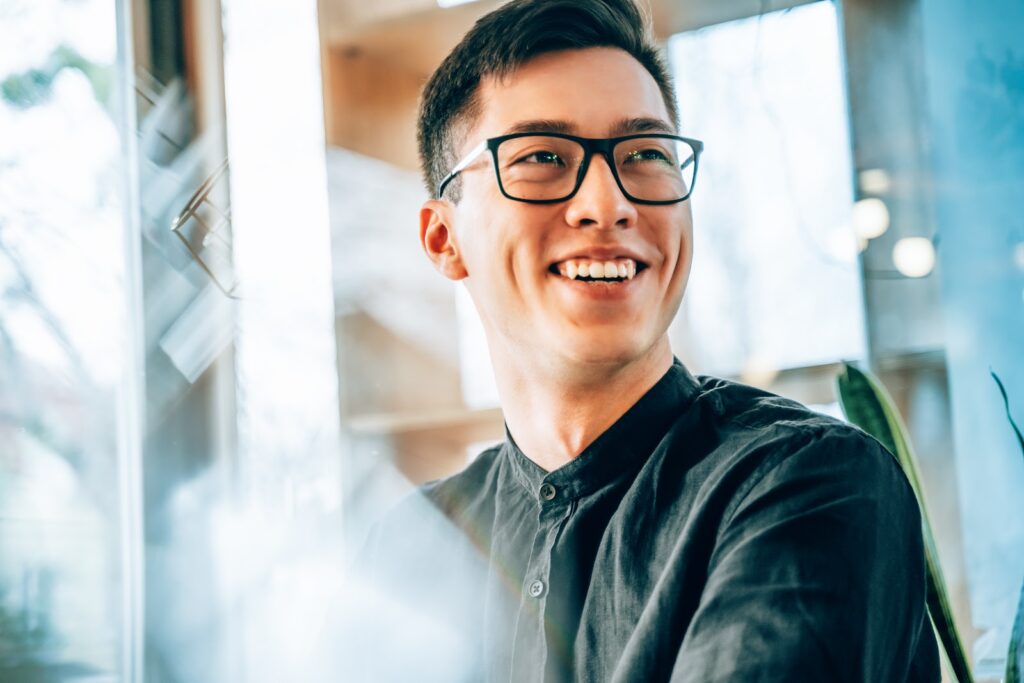 A young business person wearing glasses in an office setting smiles.