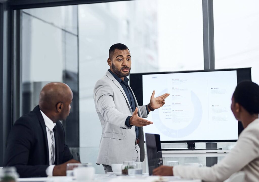 A master's of engineering management graduate in business attire delivers a presentation to colleagues in a modern office boardroom.