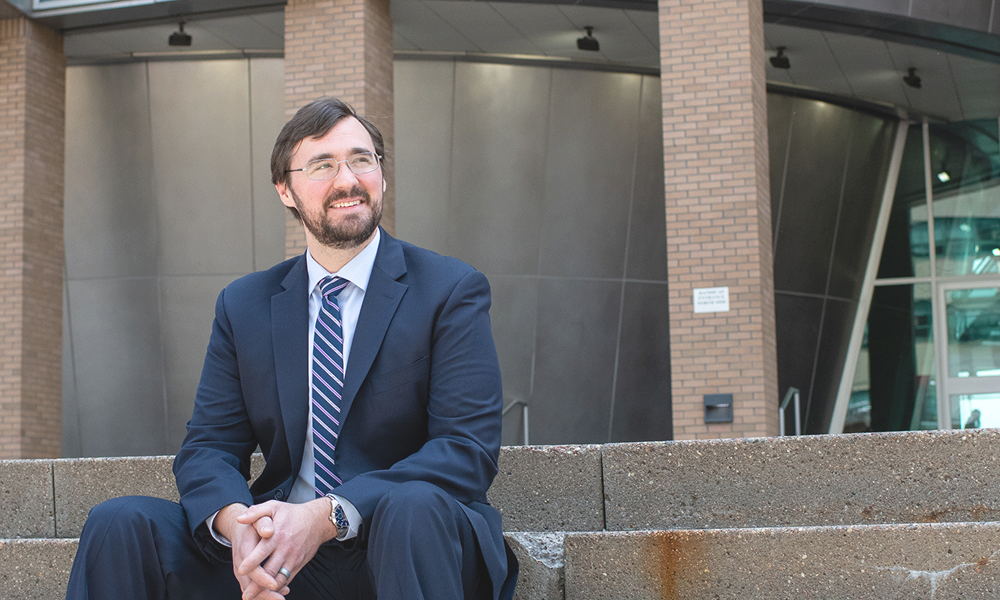 A man in a business suit, sitting and smiling.