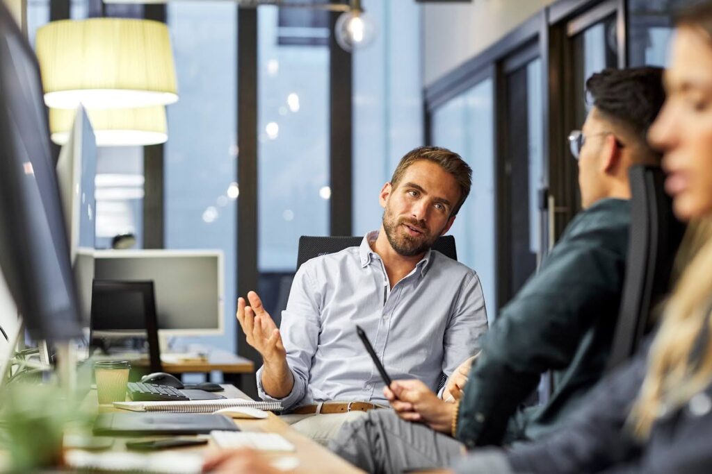 Business people sitting at computer stations face one another and converse.