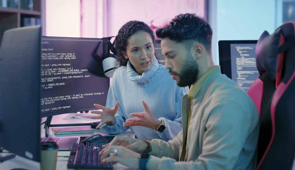 A young professional, directed by a peer, works at a multiscreen computer station; programming script is visible on several screens.