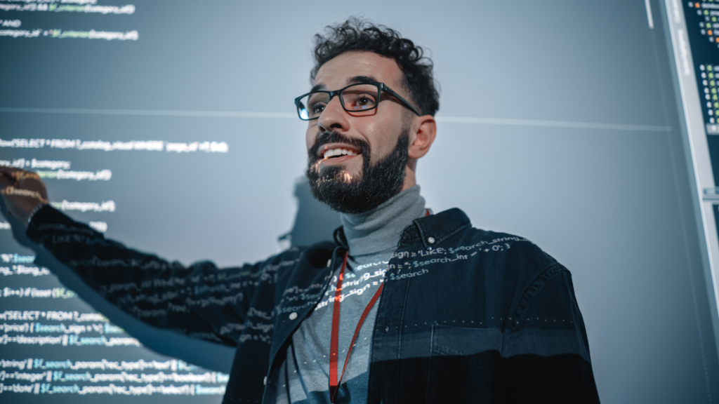 A computer science student demonstrates his work at a large screen projecting code.