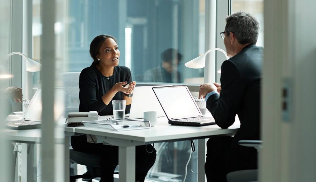 In a modern office space, two business people, each sitting at a desk with an open laptop, converse.