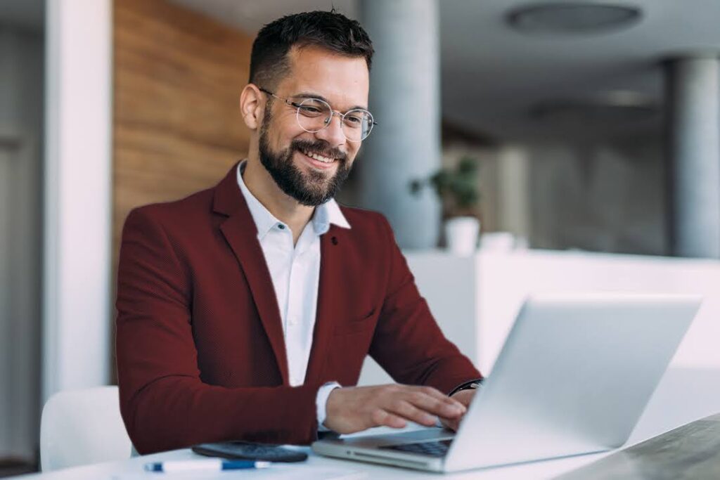 A smiling man in a red blazer works at a laptop in a modern, bright office space.